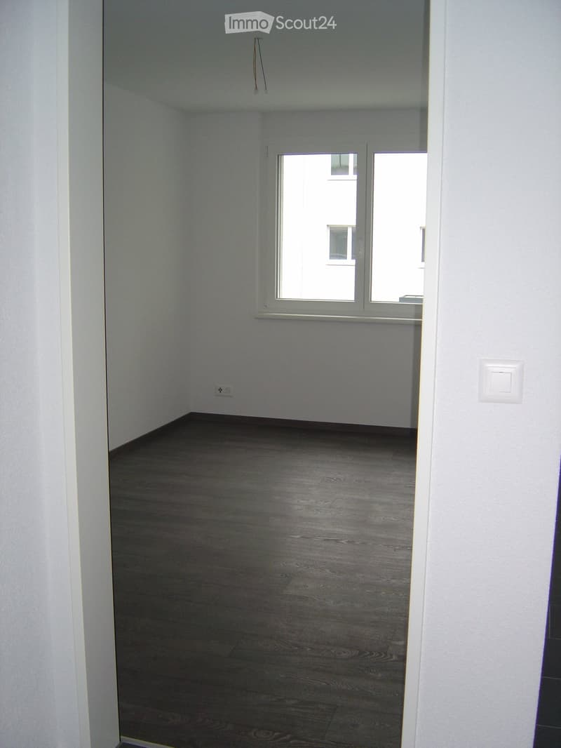 Empty room with wooden flooring and white walls, featuring a large window with a view of another building.