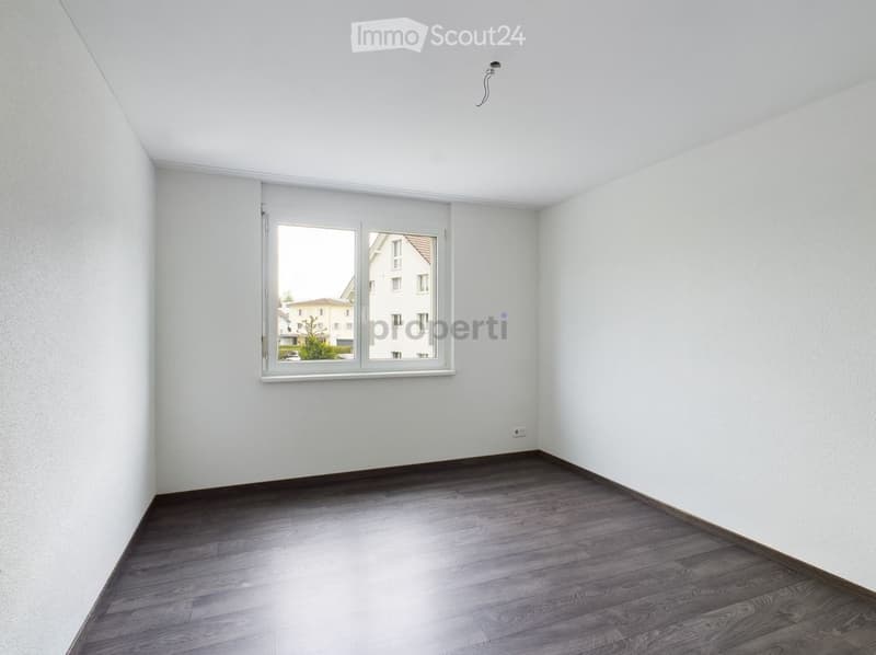 Empty room with light-colored walls, wooden flooring, and a large window offering a view of neighboring buildings.