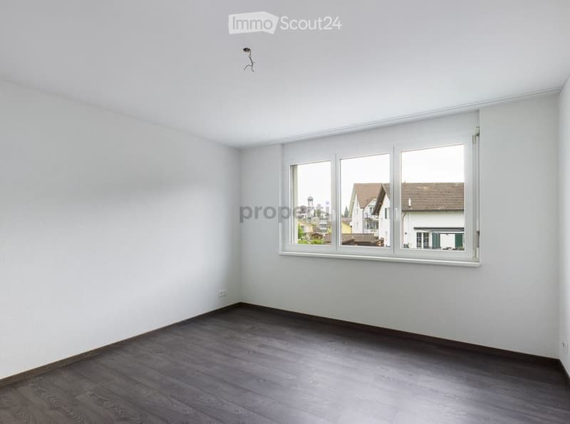 Empty room with light-colored walls, dark wooden flooring, and large windows showing a view of neighboring houses.