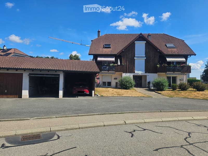 Two-story house with a tiled roof, garage, and balcony. The house features a mix of wood and white exterior walls, with a well-maintained garden and driveway.