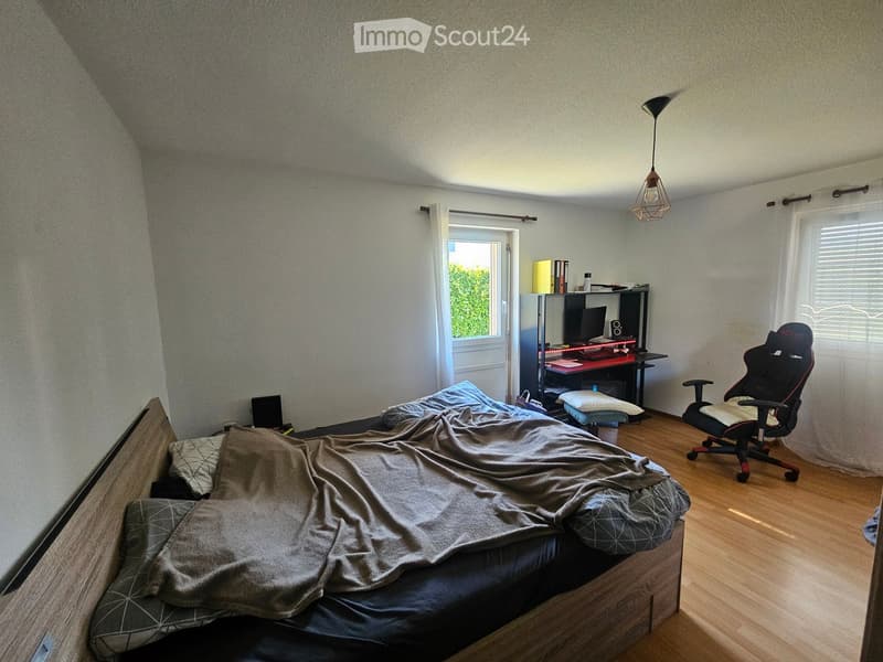 Cozy bedroom with a wooden bed, desk, chair, and a hanging lamp. The room features large windows and a door leading to an outdoor area.