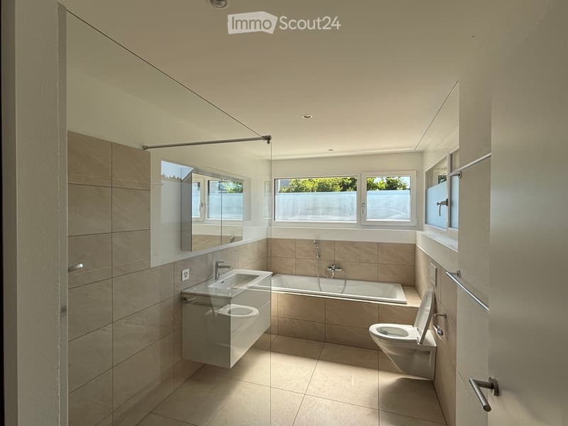 Modern bathroom with beige tiles, a large mirror, a sink, a bathtub, and a toilet. The space is well-lit with natural light from multiple windows.