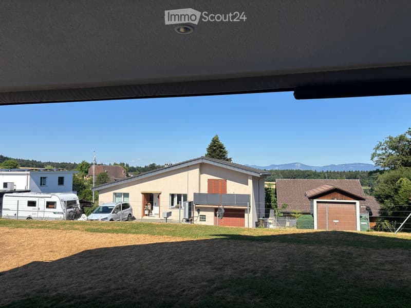 Single-story house with beige walls, red shutters, garage, and balcony. Surrounded by a grassy yard with a camper van and car parked nearby.