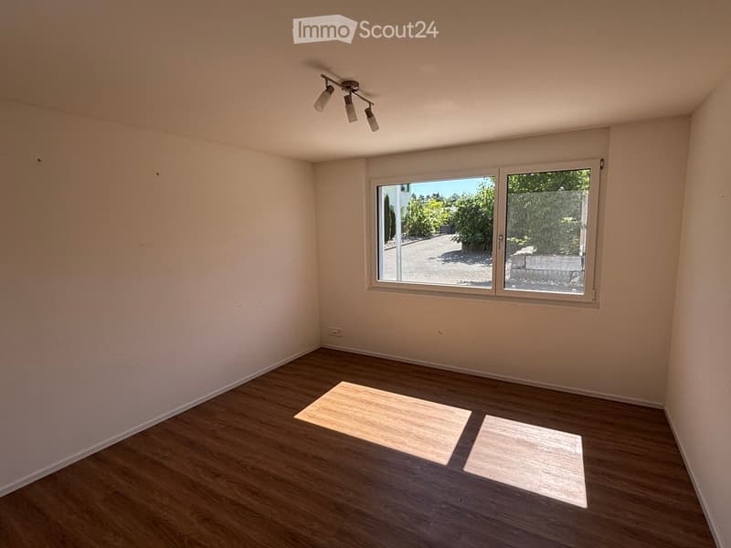 Empty room with wooden flooring, light-colored walls, and a large window offering a view of greenery outside. The room includes a ceiling light fixture and two small wooden mats placed on the floor.
