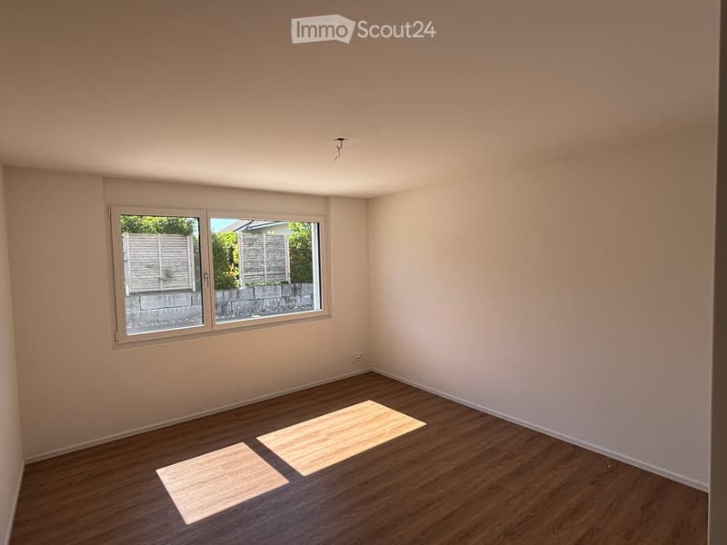Empty room with wooden flooring, light-colored walls, and a large window showing a view of a garden area with a fence and greenery.