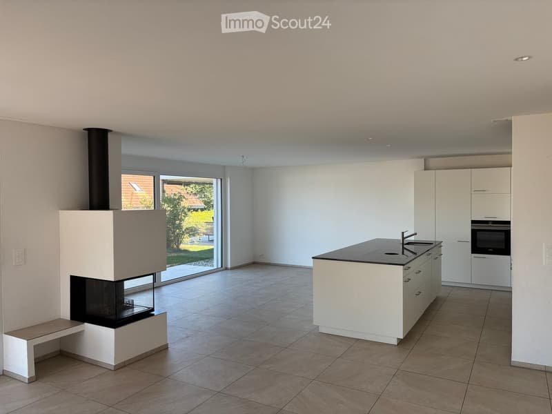 Modern kitchen with white cabinets, a central island, built-in oven, and a fireplace with a bench nearby. Large windows provide natural light and a view of the garden.