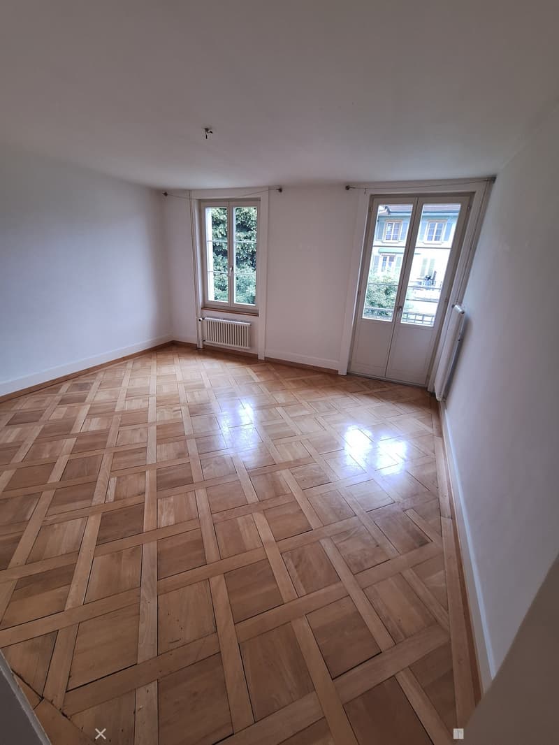 Empty room with wooden herringbone flooring, white walls, and large windows with white frames. Radiators are visible beneath the windows.