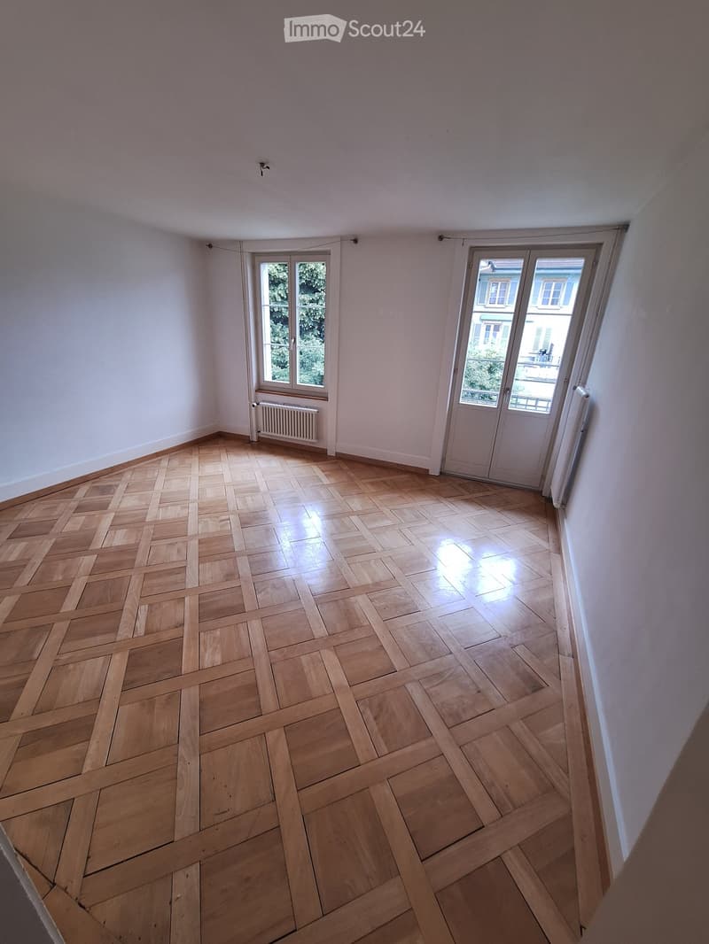 Empty room with wooden herringbone flooring, white walls, and large windows with white frames. Radiators are visible beneath the windows.