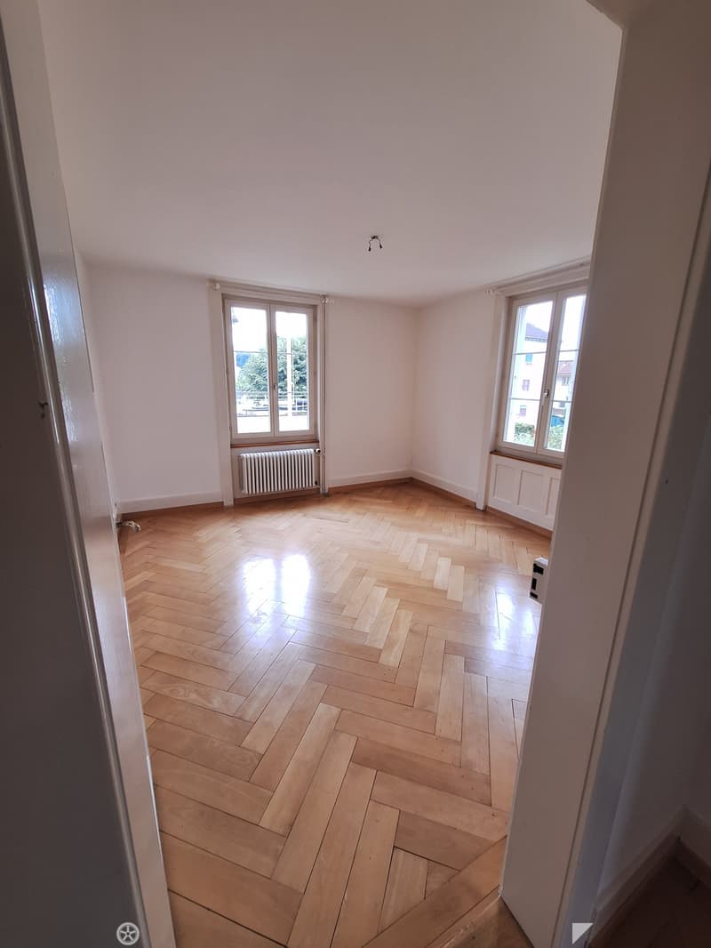 Empty room with wooden herringbone flooring, white walls, and large windows with radiators beneath them.
