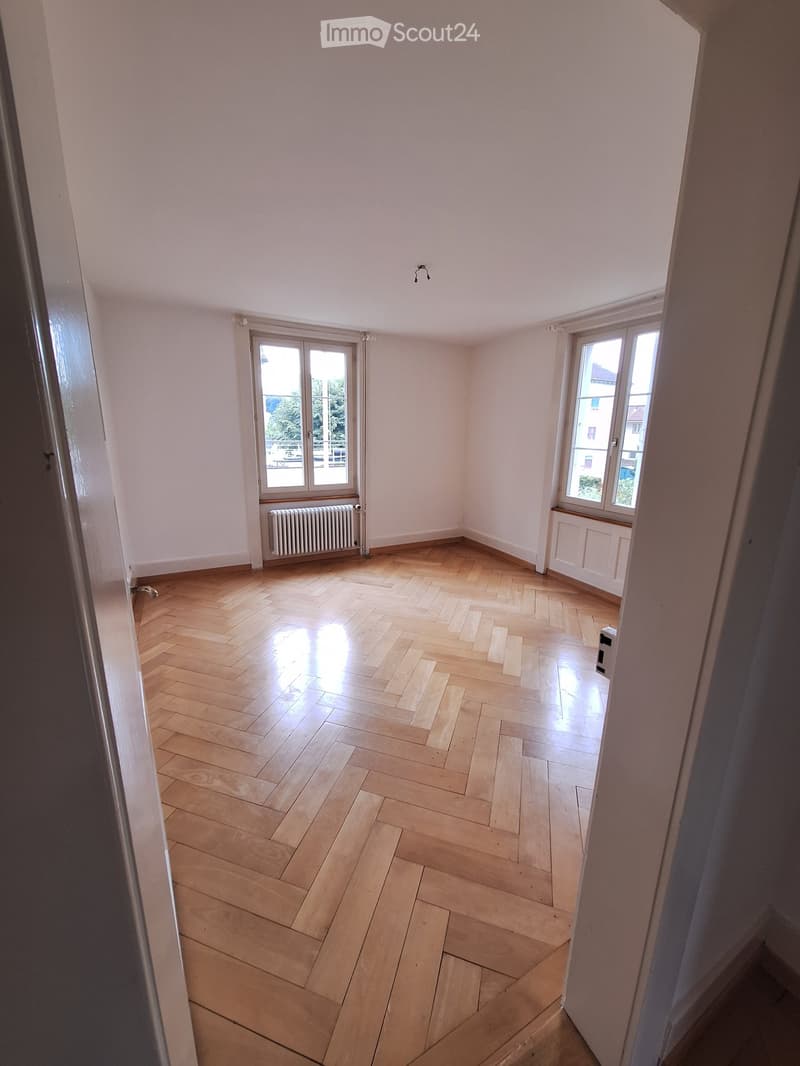 Empty room with wooden herringbone flooring, white walls, and large windows with radiators beneath them.