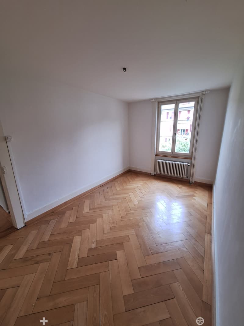 Empty room with light wooden herringbone flooring, white walls, and a window with a radiator below it.