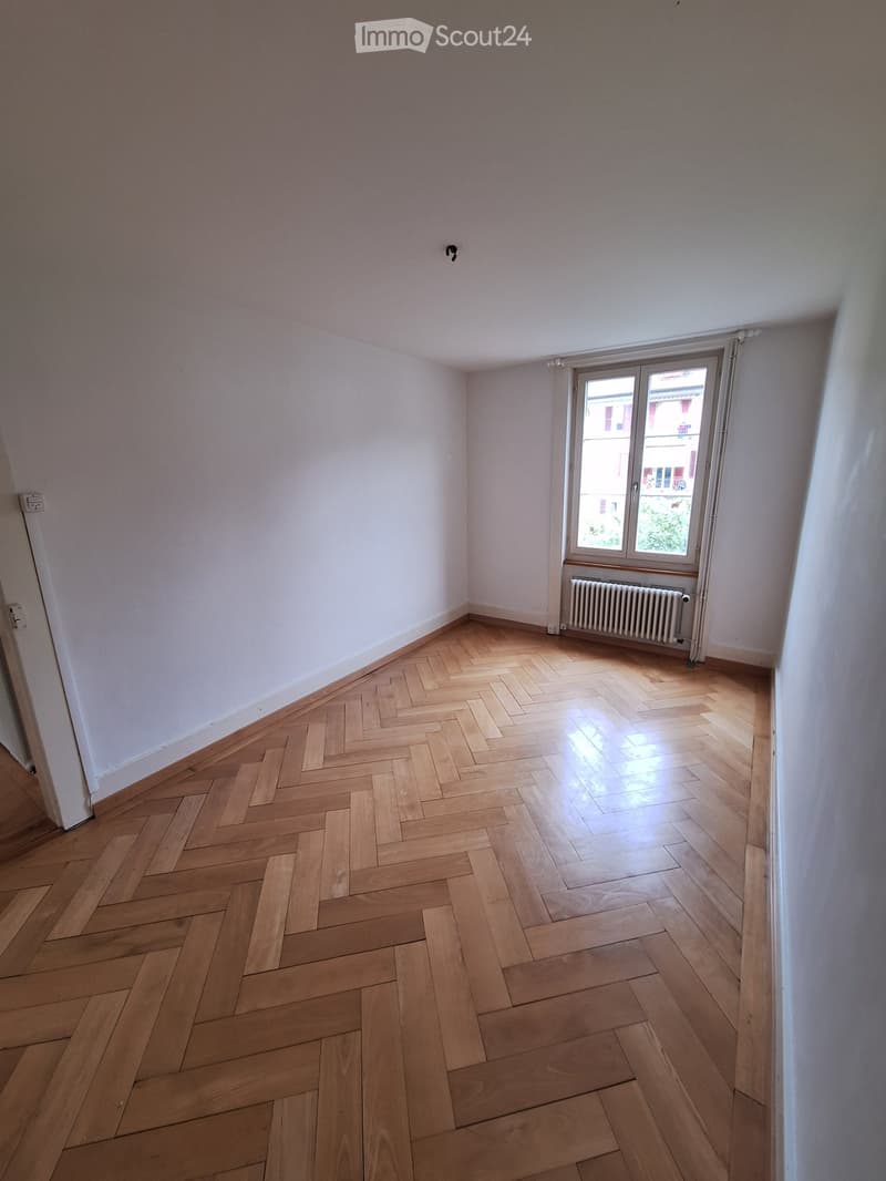 Empty room with light wooden herringbone flooring, white walls, and a window with a radiator below it.