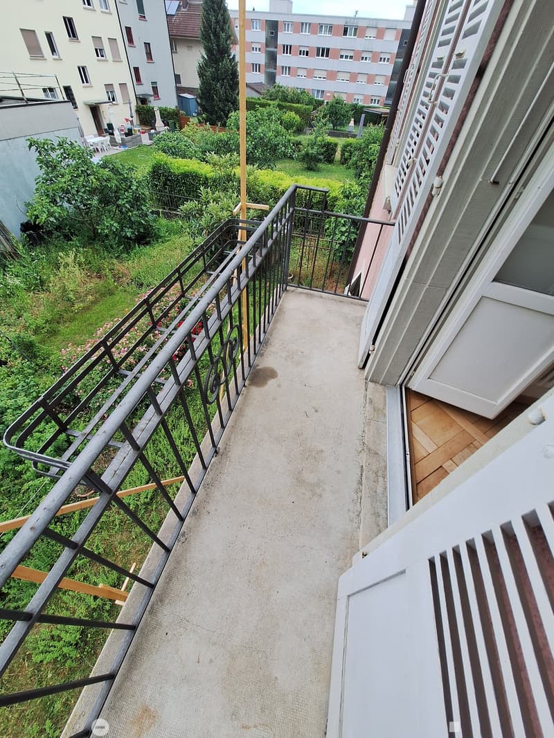 Balcony with metal railing, concrete floor, and view of a green garden area with bushes and trees.