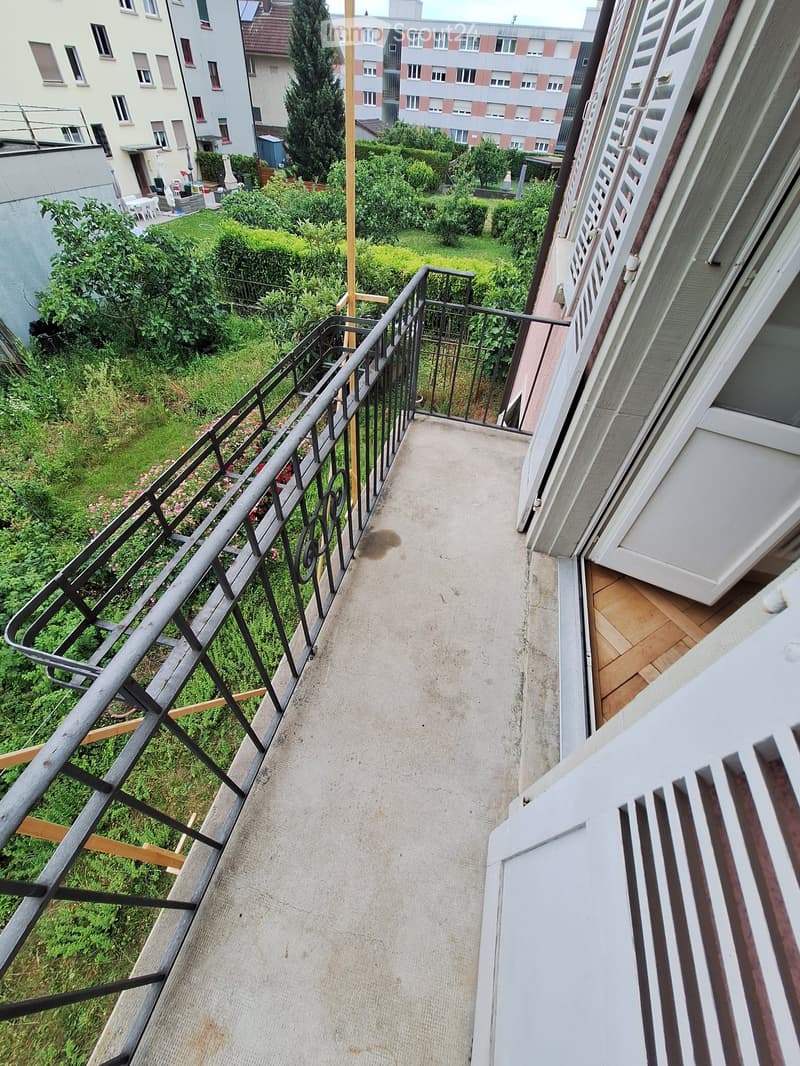 Balcony with metal railing, concrete floor, and view of a green garden area with bushes and trees.