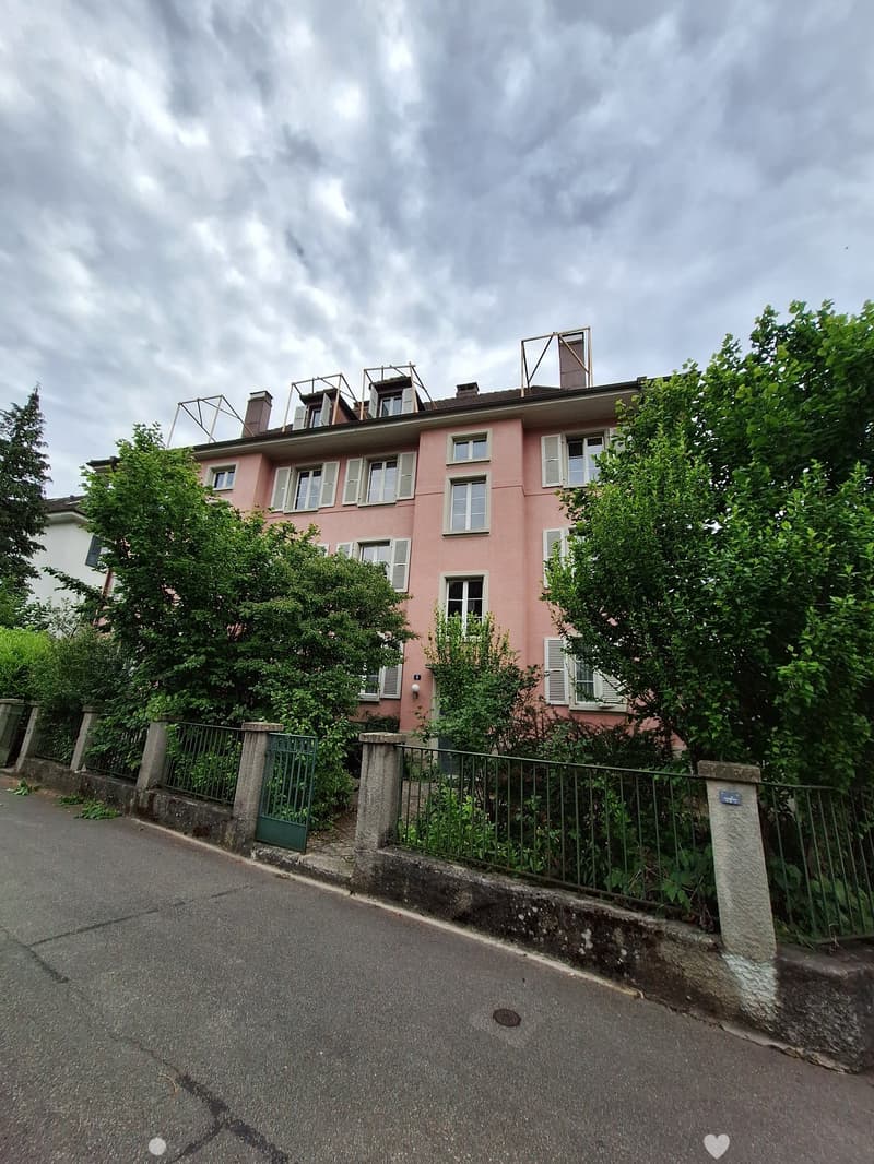 Pink two-story house with multiple windows, shutters, and a small rooftop terrace, surrounded by greenery and a metal fence.