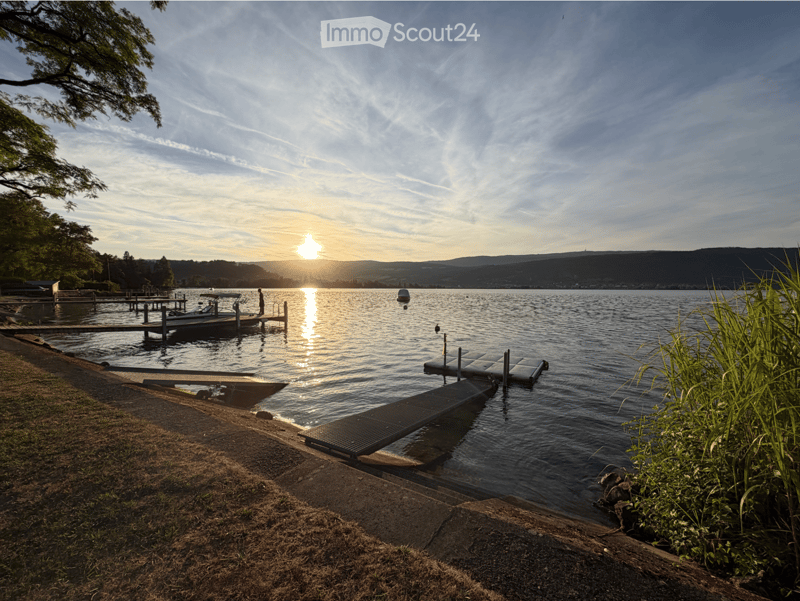 A serene lakeside scene at sunset with a dock, a boat, and a calm lake reflecting the sky and surrounding trees.