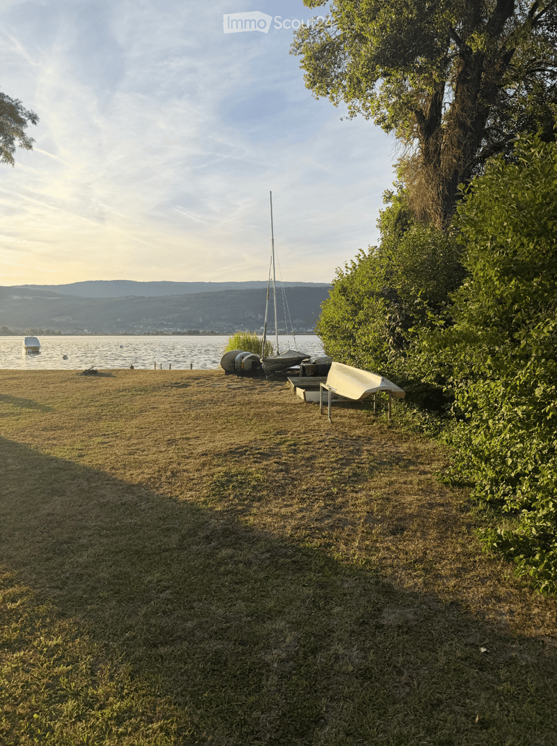 Lush green garden area near a lake with a sailboat, lounge chair, and small table.