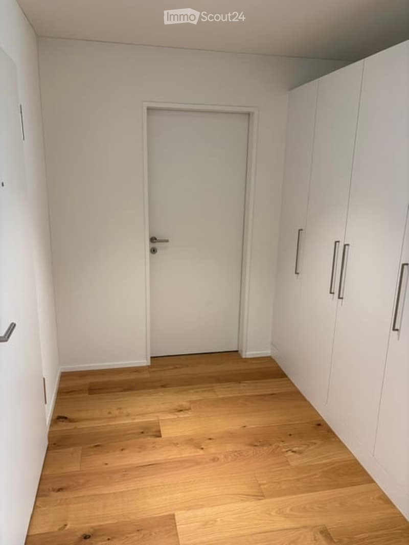 Empty room with light-colored walls, wooden flooring, and built-in white wardrobes on one side. A closed door is visible in the center.