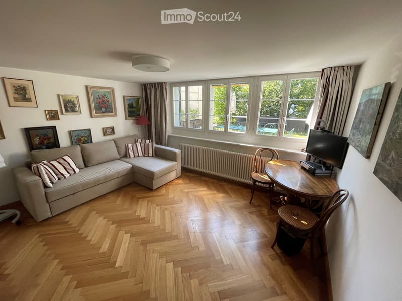 Living room with light wooden herringbone flooring, large windows with curtains, a cream sectional sofa with striped cushions, a wooden coffee table with a TV, and several framed artworks on the walls.