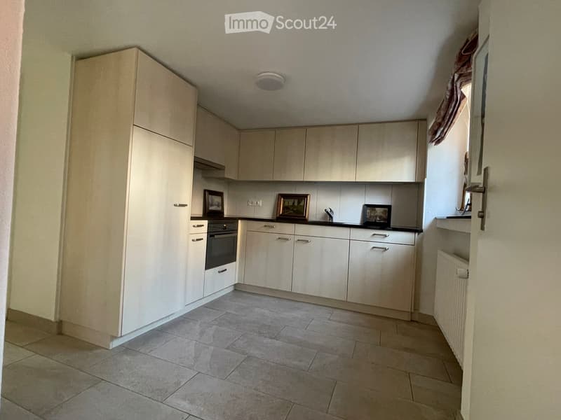 Modern kitchen with light-colored cabinets, tiled floor, and an oven. There are framed pictures on the counter and a small window with a curtain.