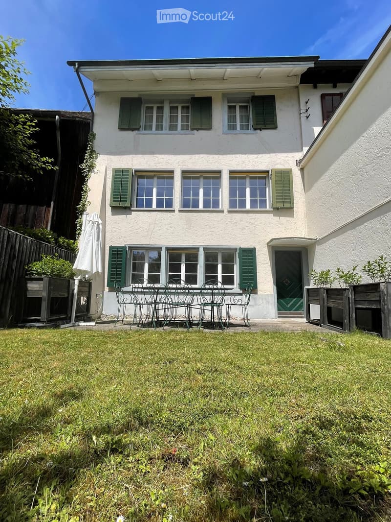 A two-story house with white walls and green shutters, featuring a small outdoor seating area with a table and chairs on a grassy lawn.