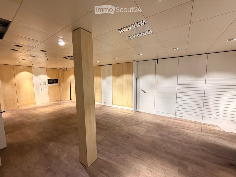 Empty room with wooden flooring, white walls, and a central wooden pillar. The space features recessed lighting and a large white panel wall on one side.