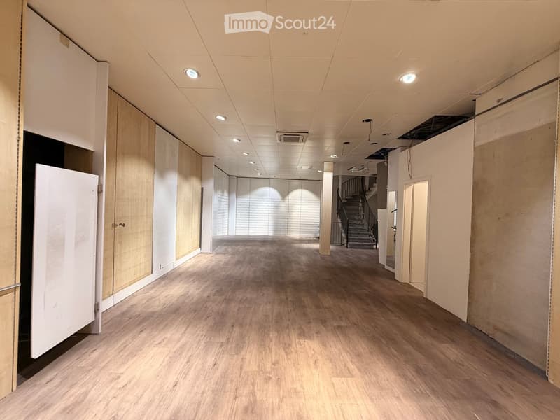 Empty hallway with wooden flooring, white walls, and recessed lighting. Features a staircase leading upwards and several closed doors along the sides.