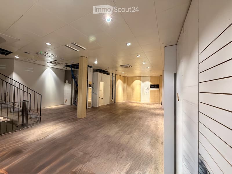 Empty hallway with wooden flooring, white walls, and modern lighting fixtures. Features a staircase on the left and a wooden pillar in the center.