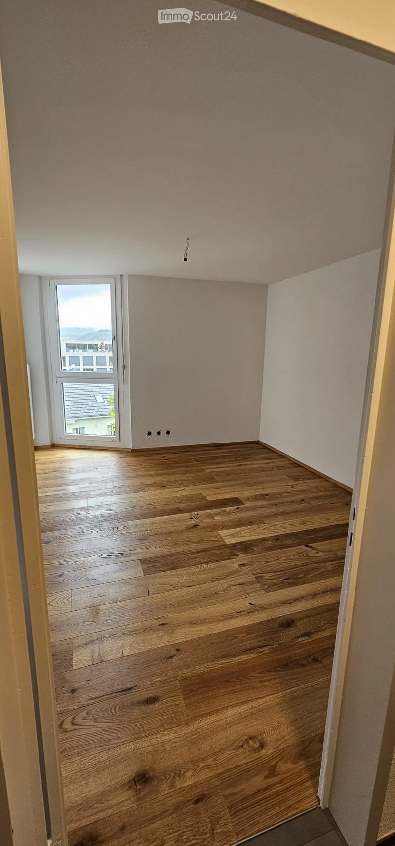 Empty room with wooden flooring and white walls, featuring a large window with a view of the outside.