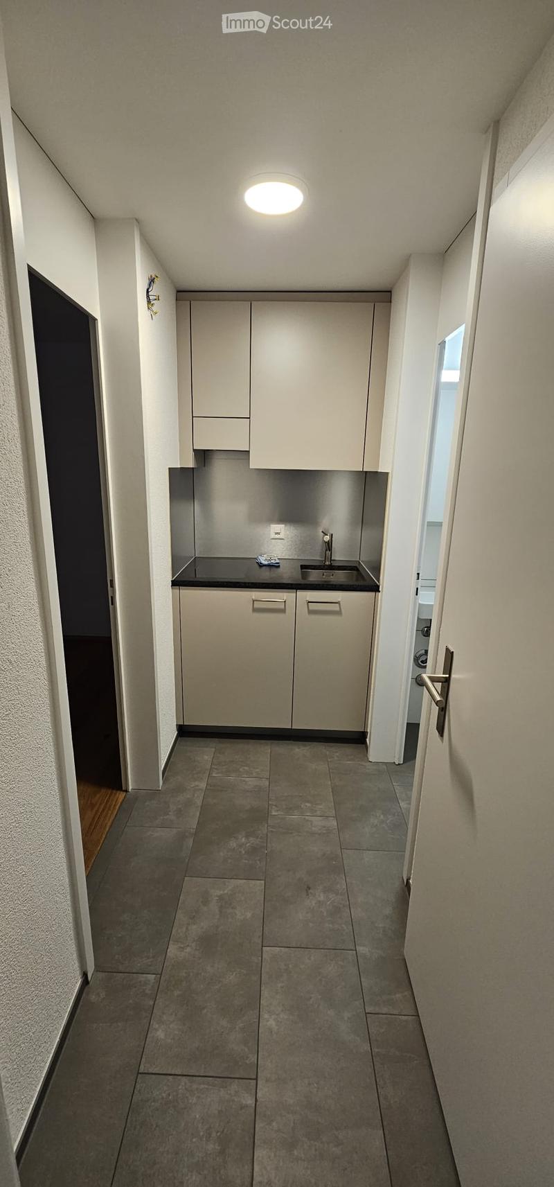 A narrow hallway with a small kitchenette area featuring cabinets, a sink, and a countertop. The floor is tiled, and the walls are painted white.
