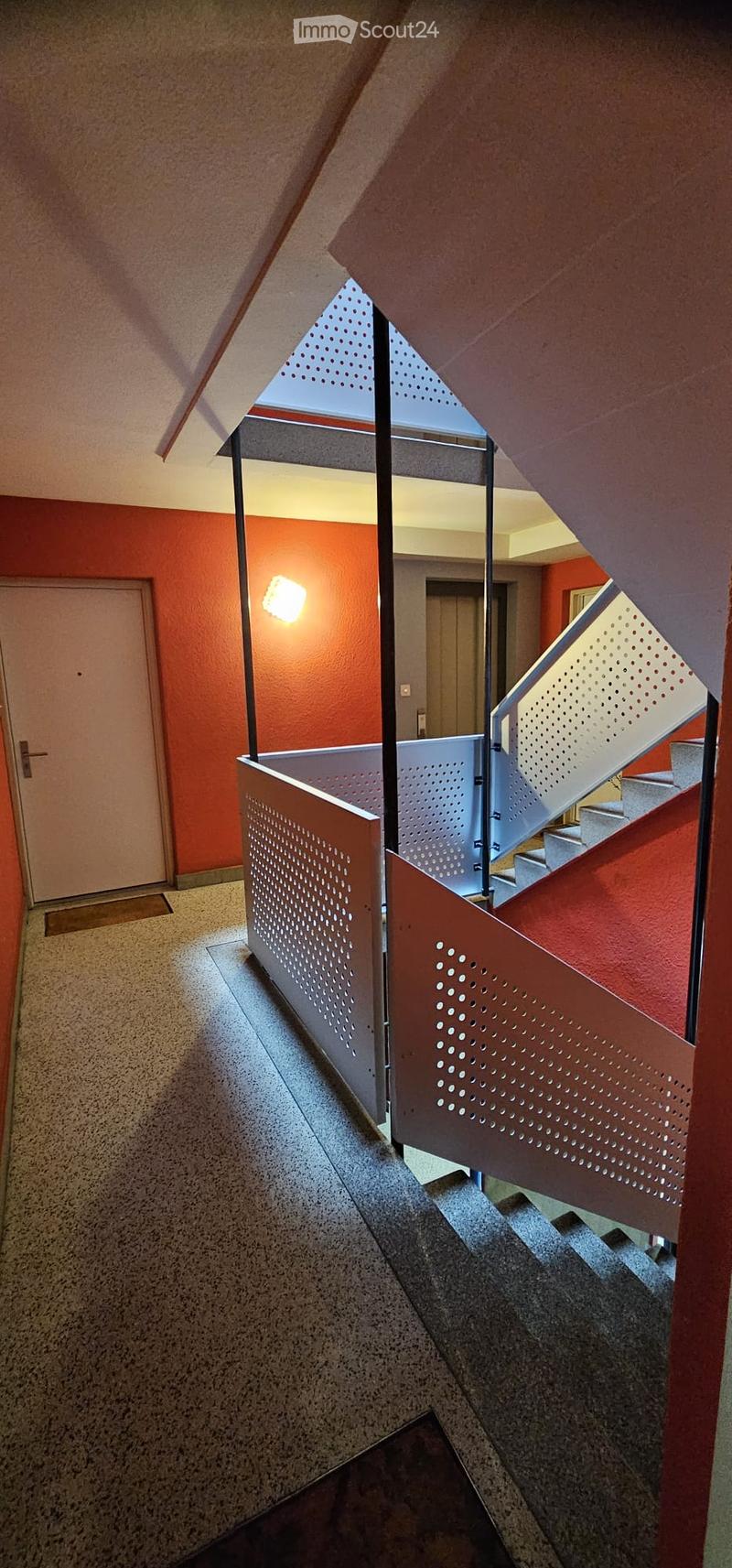 Modern hallway with red walls, a staircase with perforated metal railings, and a bright light fixture on the wall.