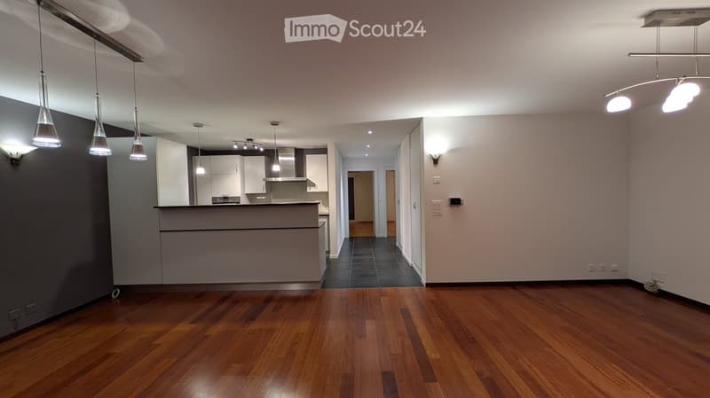 Open-concept unfurnished room with hardwood flooring, modern lighting fixtures, and a small kitchen area visible in the background.