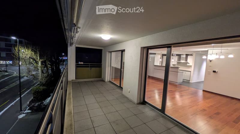 Balcony with tiled floor, metal railing, and sliding glass doors leading to a modern kitchen with white cabinets and wooden flooring.