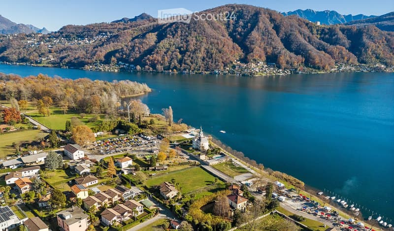 Aerial view of a lakeside town with houses, a parking lot, and a church, surrounded by mountains and a lake.