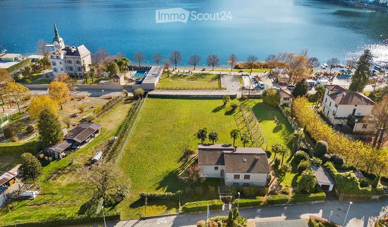 Aerial view of a large, well-maintained lawn area surrounded by trees and buildings, with a scenic lake view in the background.