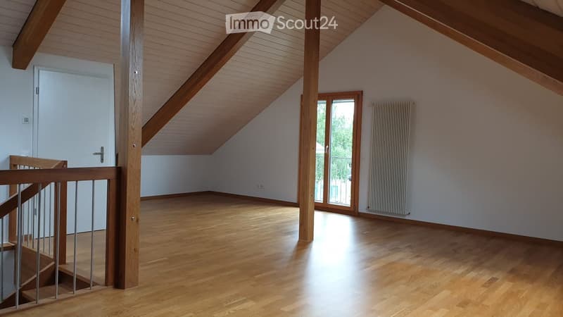 Bright, empty attic room with wooden floors, white walls, exposed wooden beams, and a staircase leading down. Features a large window and a radiator on one wall.