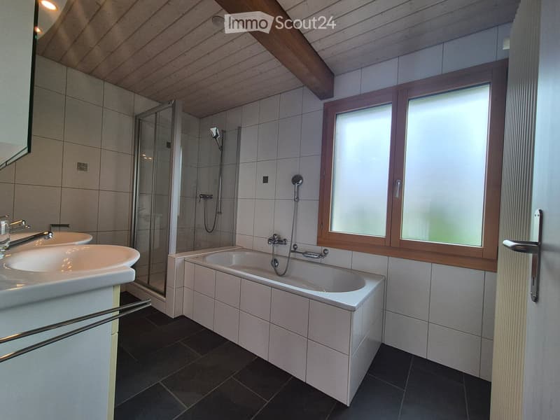 Modern bathroom with a bathtub, double sink, shower, and large window with frosted glass. The room features wooden beams on the ceiling and tiled walls.