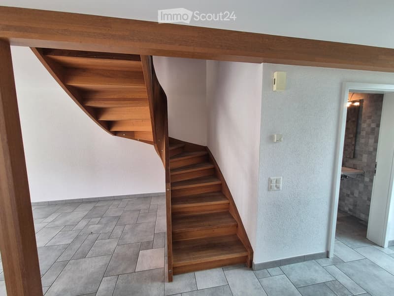 Wooden staircase with built-in shelving, light-colored walls, tiled floor, and a small adjacent room visible.