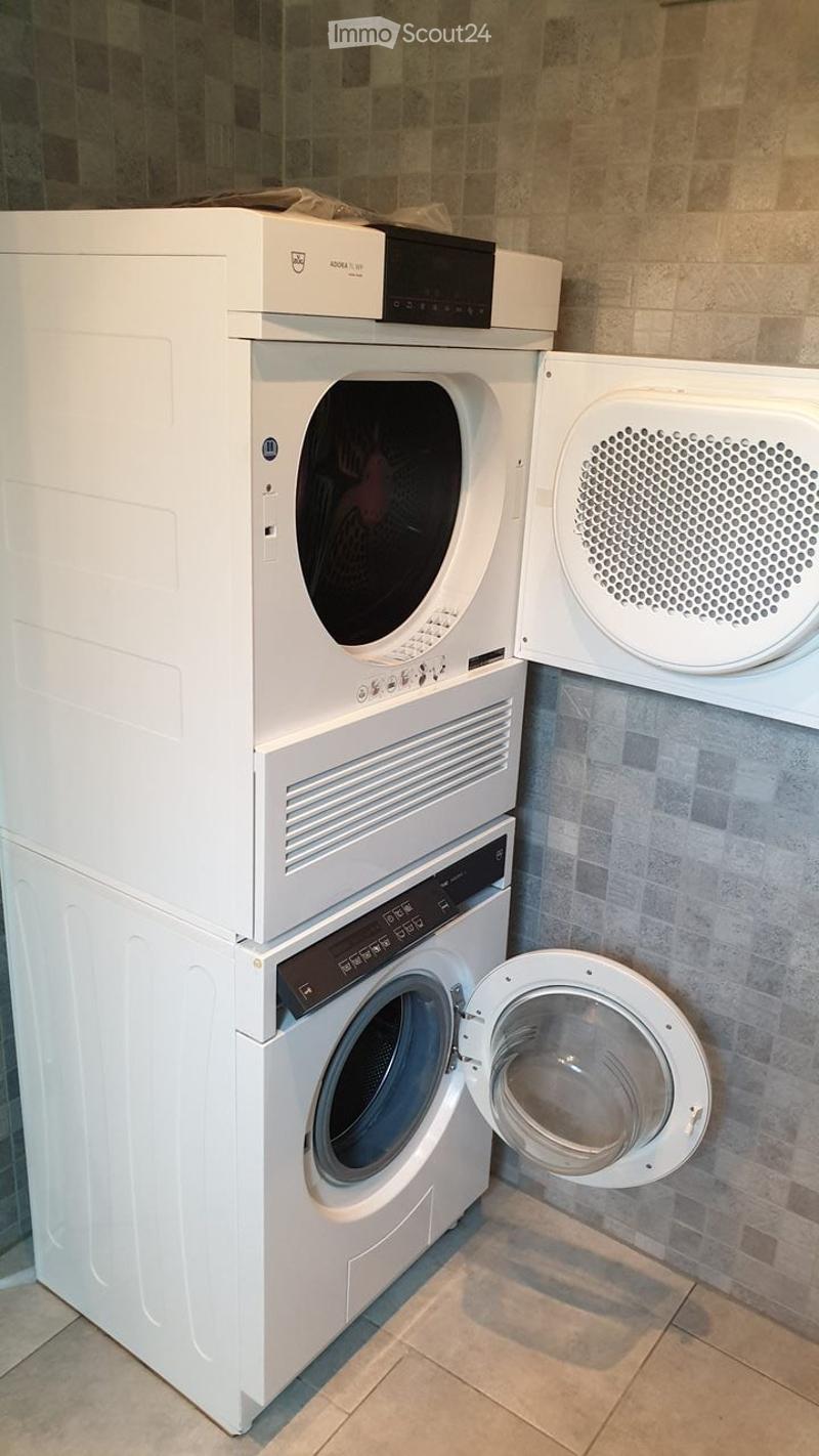 A modern bathroom with a white washing machine and dryer stacked vertically, featuring a tiled wall and tiled floor.