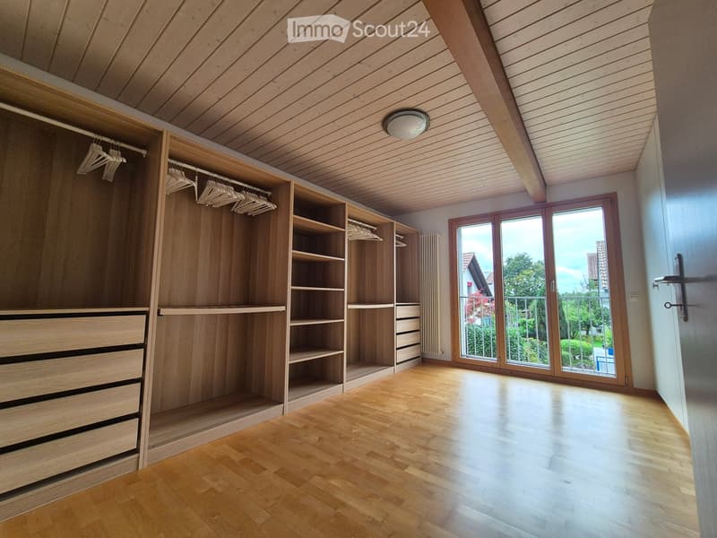 Empty room with wooden floor, built-in wardrobe with shelves and drawers, large window with view of garden, and modern lighting fixture on the ceiling.
