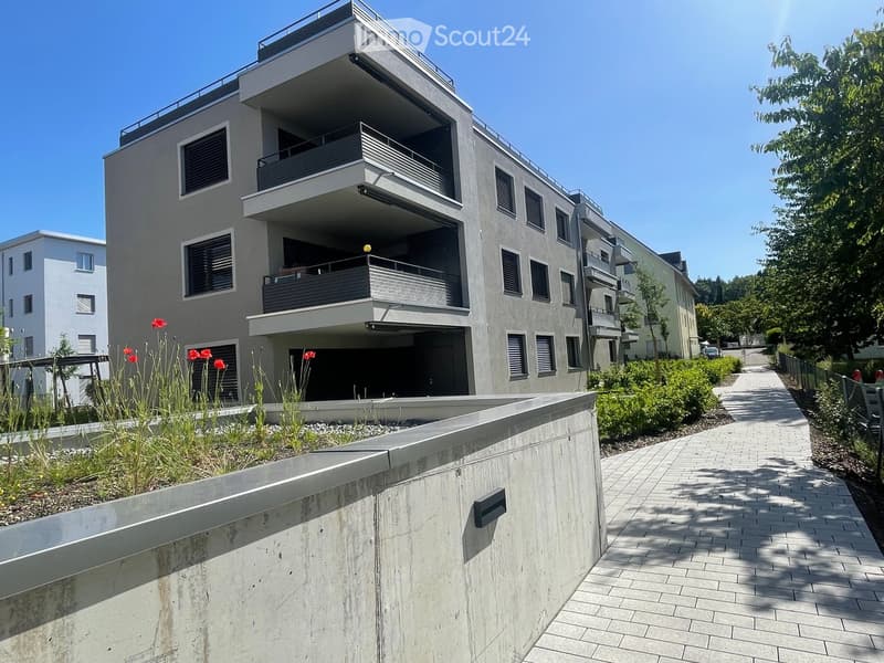multistory building, balconies, gray walls, bricked pathway, shrubs, parking area in front
