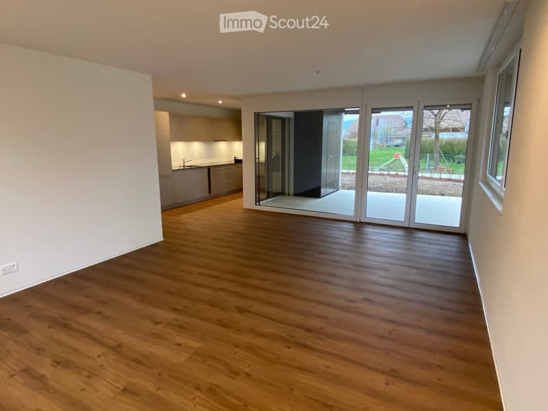 Empty room with wooden flooring, white walls, and large sliding glass doors leading to an outdoor area. The adjacent kitchen is visible in the background.