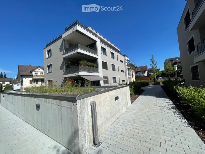 Modern multi-story residential building with balconies, surrounded by greenery and a paved walkway.