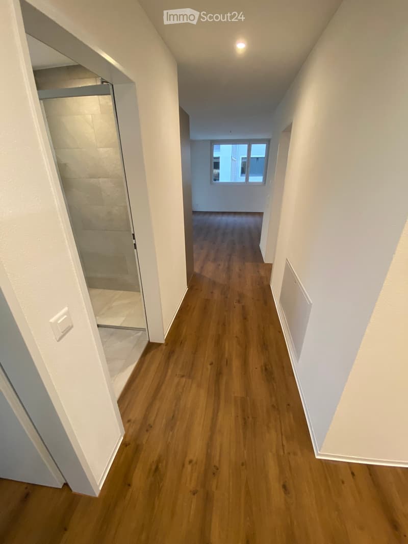Empty hallway with wooden flooring, white walls, and a window at the end. The hallway leads to an open area with a visible room on the left.