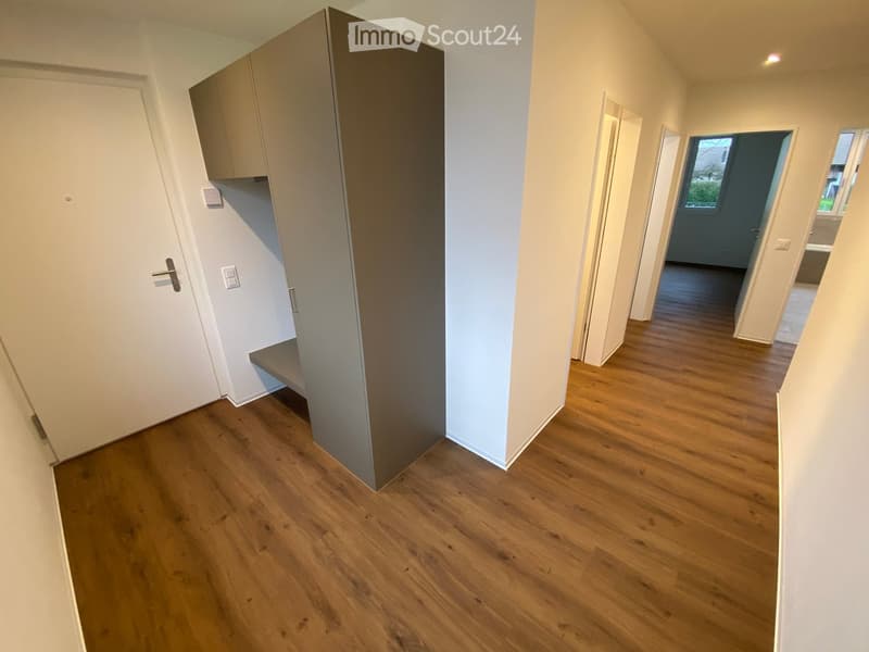 Empty hallway with wooden flooring, white walls, and several doors leading to other rooms. Natural light comes through a window at the end of the hallway.