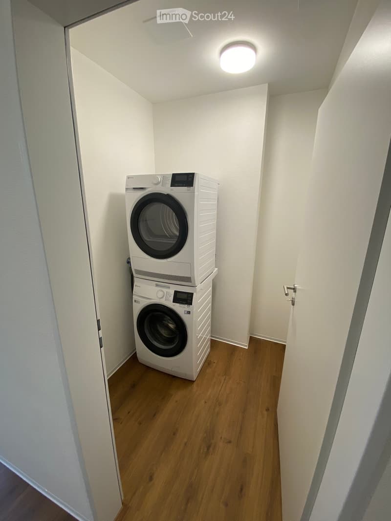 Small room with wooden flooring and white walls, containing a stacked washing machine and dryer unit.