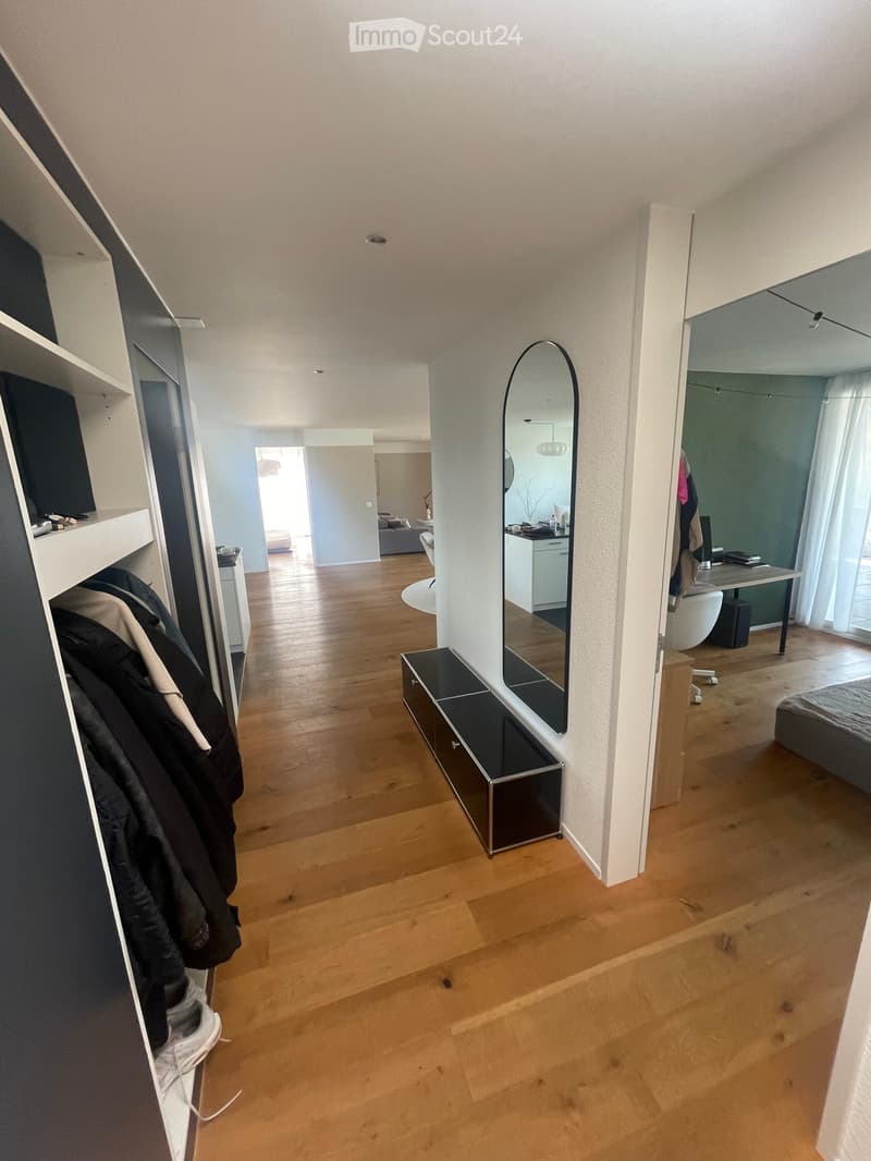 A well-lit hallway with wooden flooring, leading to a bedroom and a bathroom. Features include a built-in wardrobe, a large mirror, and a small black storage unit.