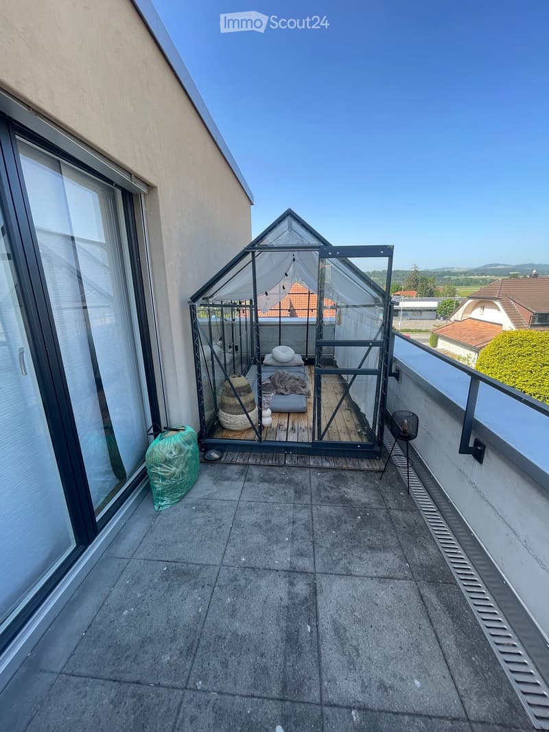 Balcony with a small enclosed seating area featuring a wooden floor, a wicker basket, a stool, and a chair. The balcony has a concrete floor and overlooks a residential area with houses and greenery.