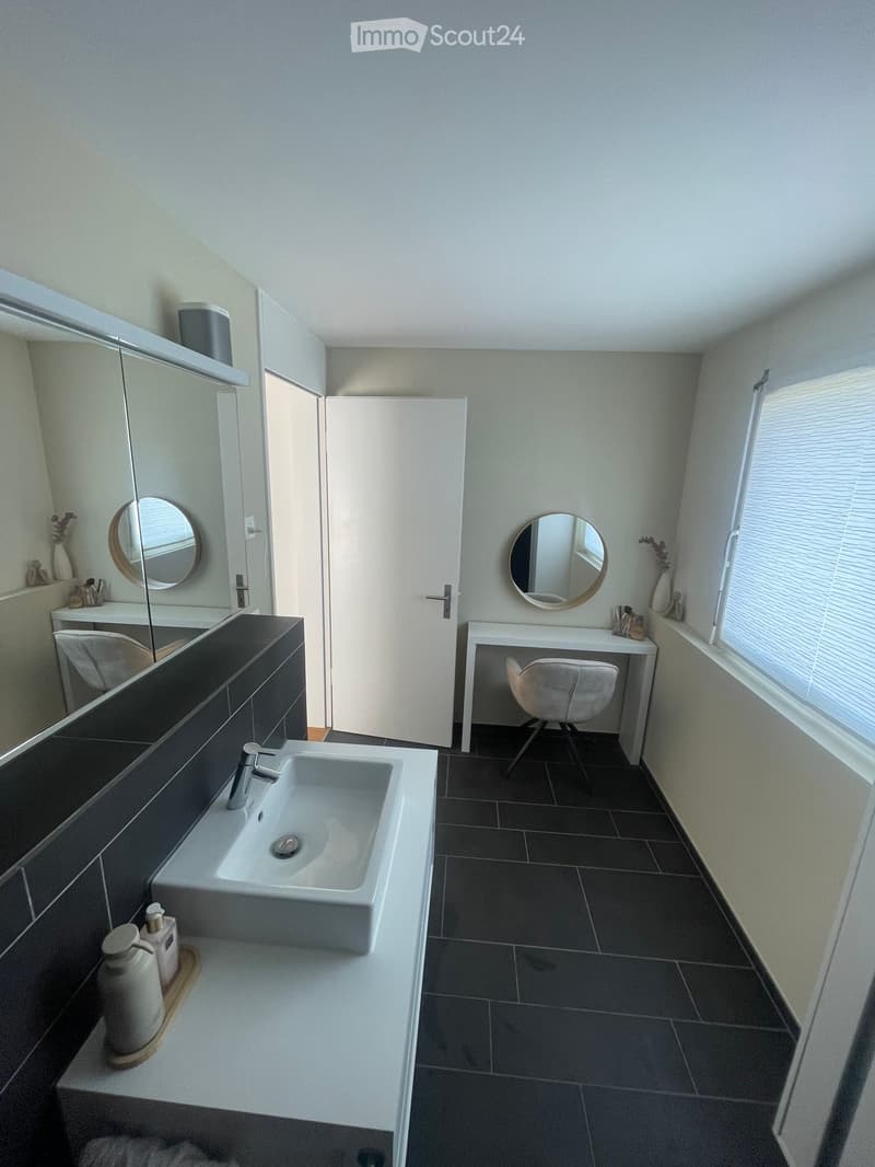 Modern bathroom with a white sink, large mirror, round wall mirrors, a small table with a chair, and decorative vases on the windowsill.