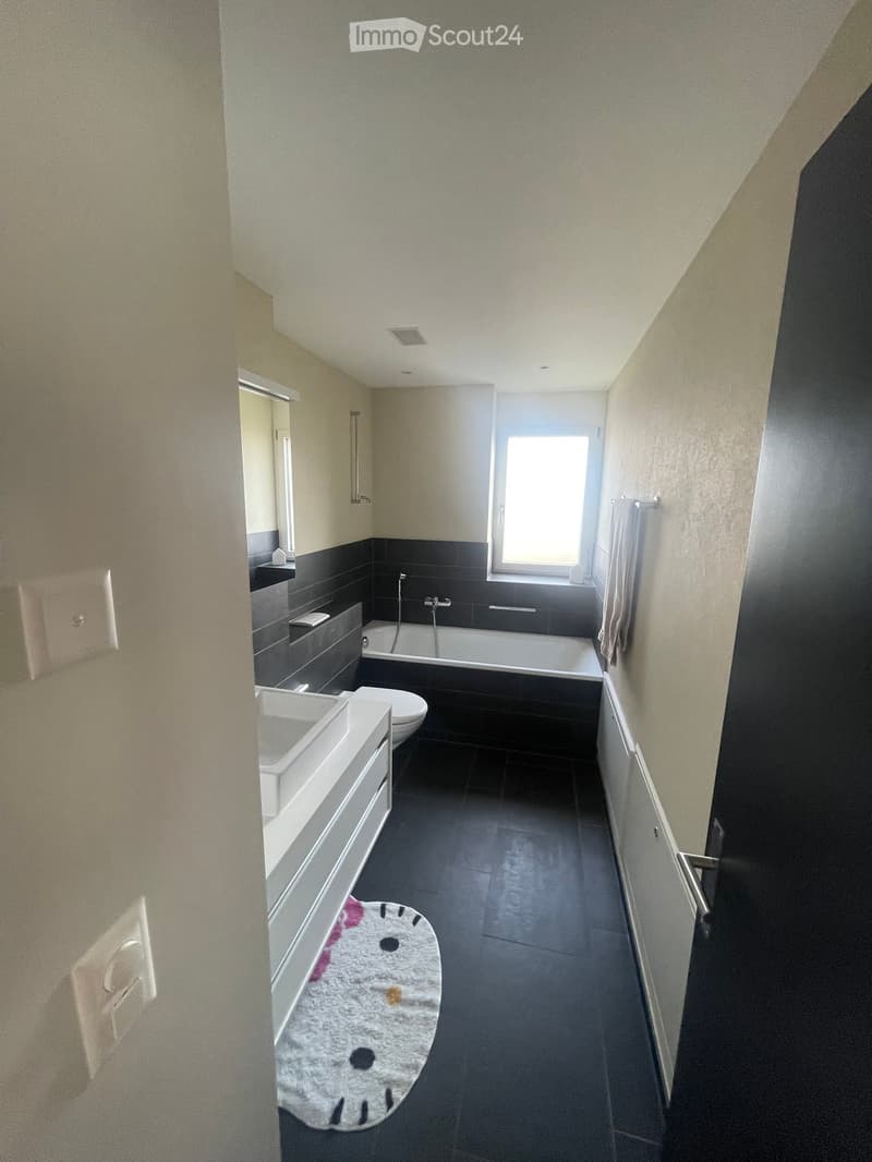 Modern bathroom with a bathtub, toilet, sink, and mirror. Features dark tiles and a small window letting in natural light.